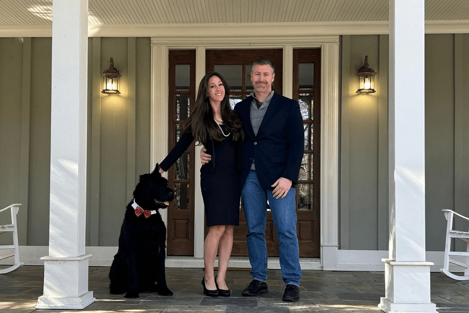 A couple, smiling, stands on a porch beside a seated black dog wearing a red bow tie. The setting is illuminated by warm porch lights, creating an inviting atmosphere.