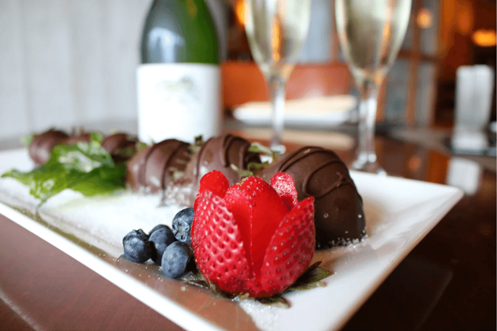 A platter of chocolate-covered strawberries and blueberries garnished with mint, with champagne flutes in the background.