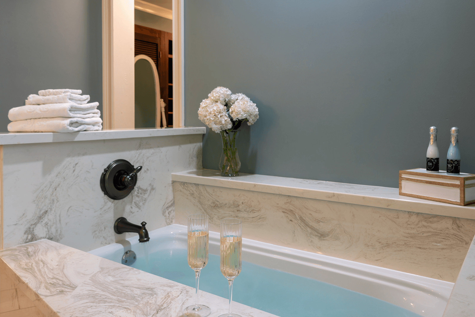 A luxurious bathroom featuring a soaking tub, folded towels, a vase of flowers, and two glasses of champagne.
