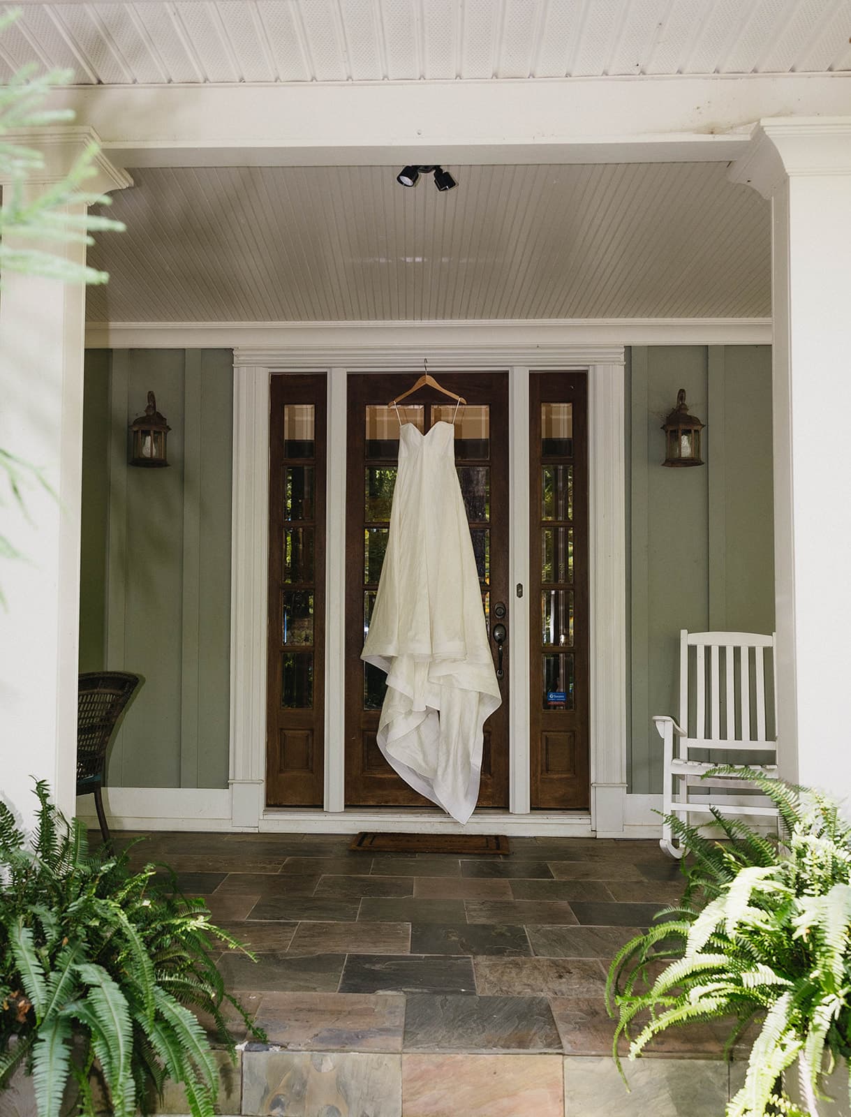 A white wedding dress hangs elegantly on a wooden door of a porch, flanked by green plants. The scene is serene, with a white rocking chair on the right.