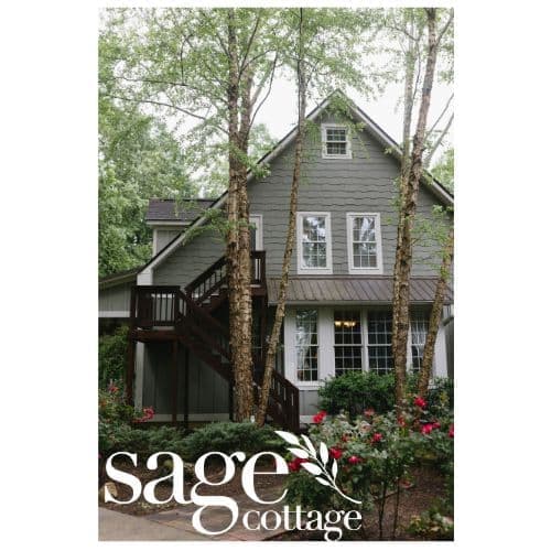A charming gray cottage surrounded by trees and colorful flowers.