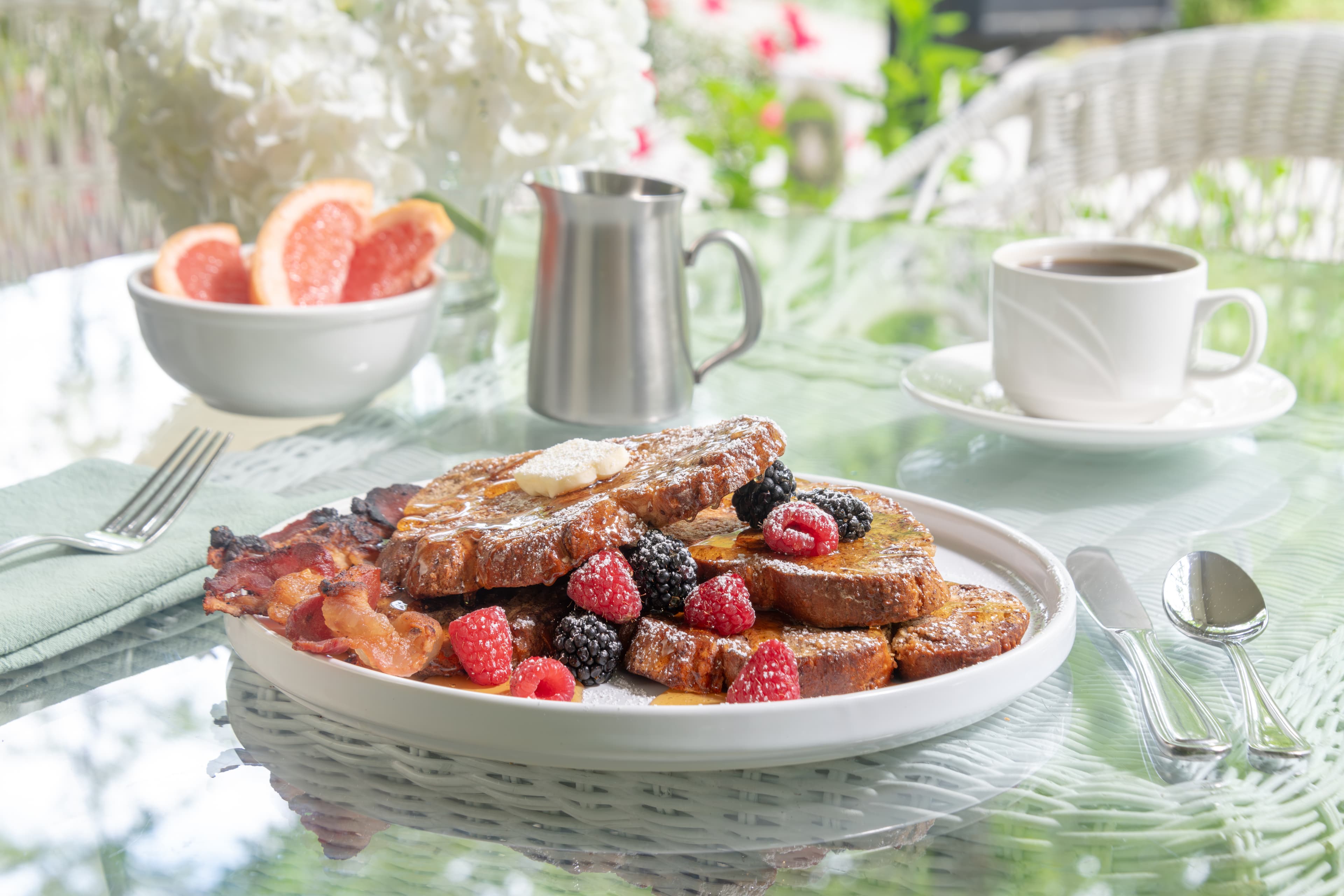 A plate of French toast topped with berries and butter, accompanied by bacon, grapefruit, a silver milk jug, and a cup of coffee on a glass table.