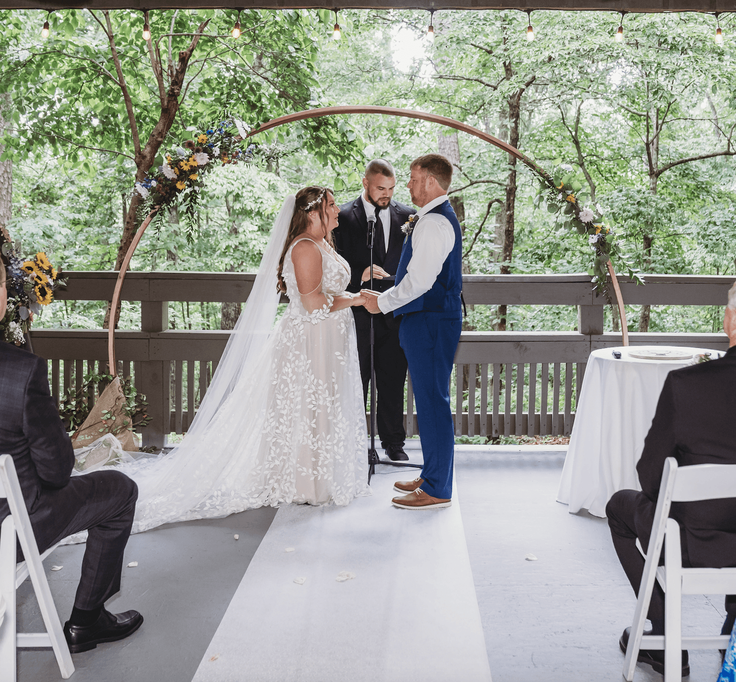 A couple exchanges vows during their wedding ceremony under a floral arch in a forest setting. A couple exchanges vows during their wedding ceremony under a floral arch in a forest setting.