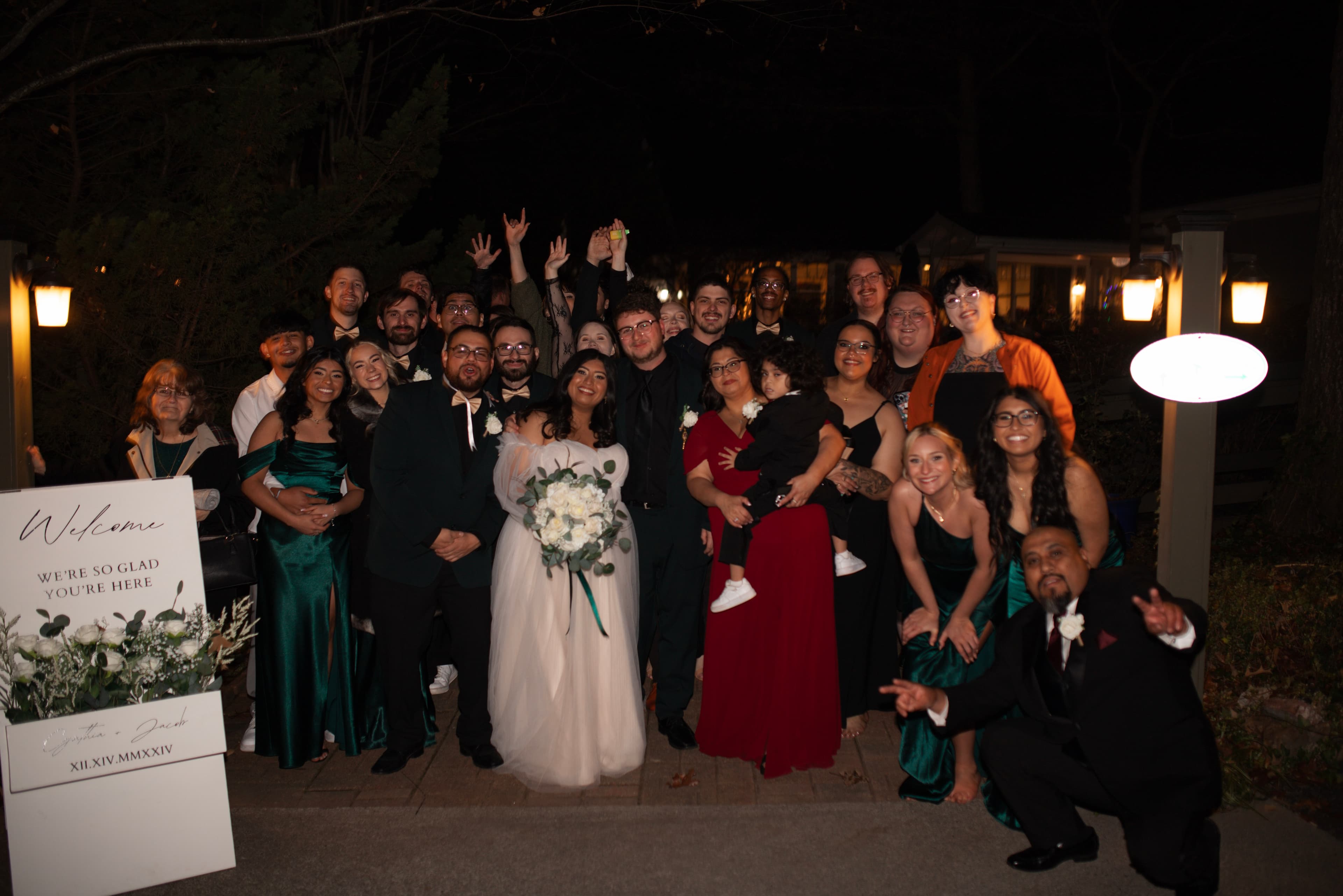 A joyful wedding party gathers for a group photo outside at night.