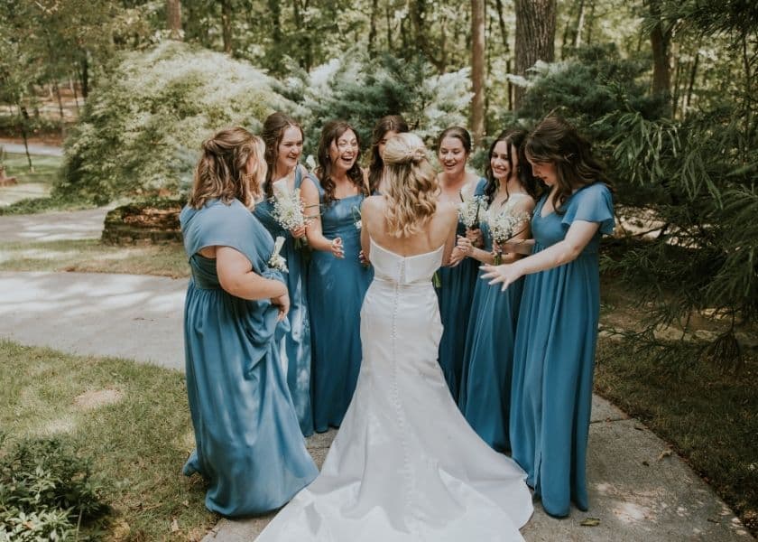 A bride in a white gown is surrounded by seven bridesmaids in blue dresses, joyfully celebrating in a garden setting.