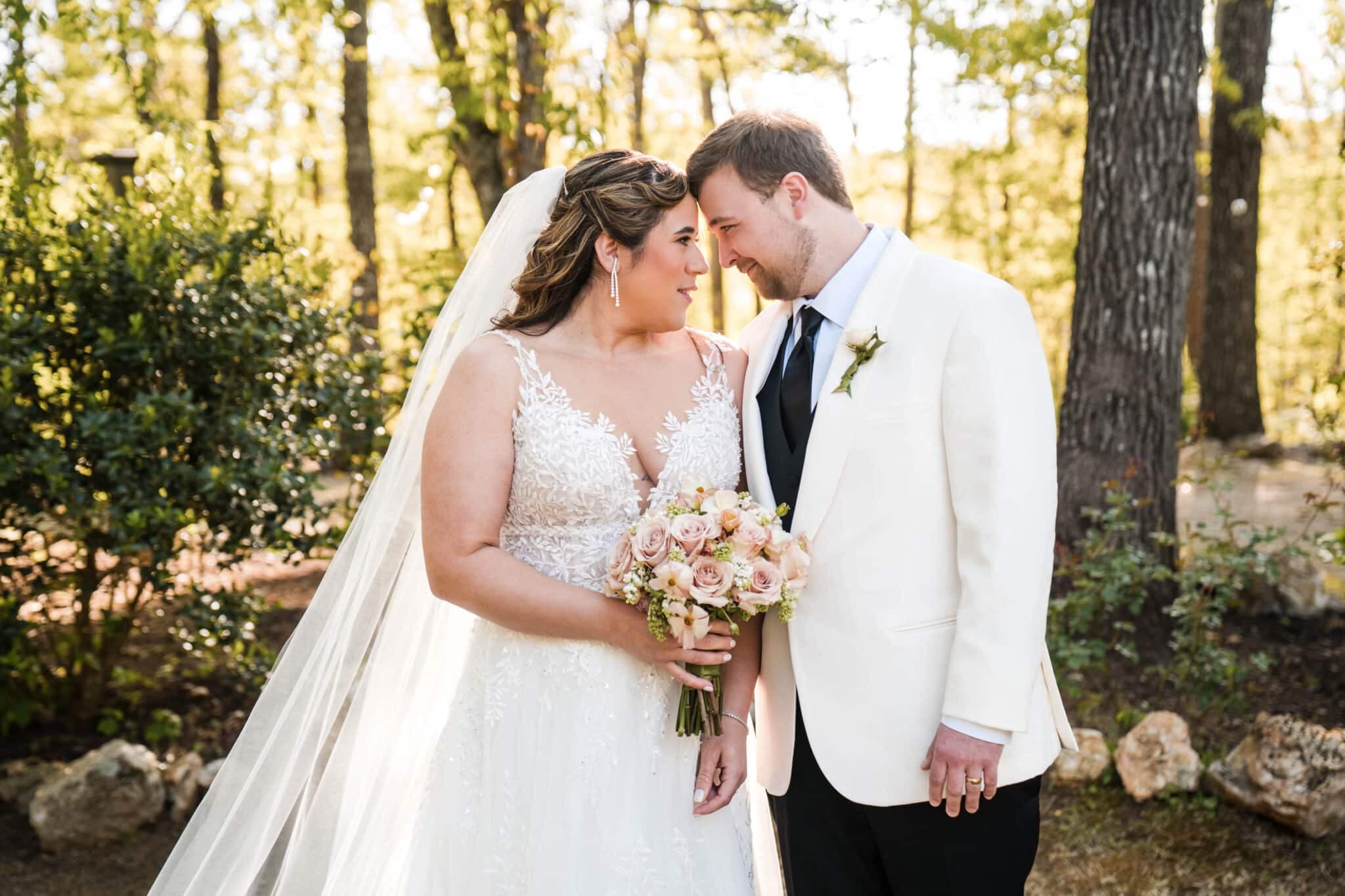 A bride and groom lean in close to each other, smiling in a woodland setting. A bride and groom lean in close to each other, smiling in a woodland setting.