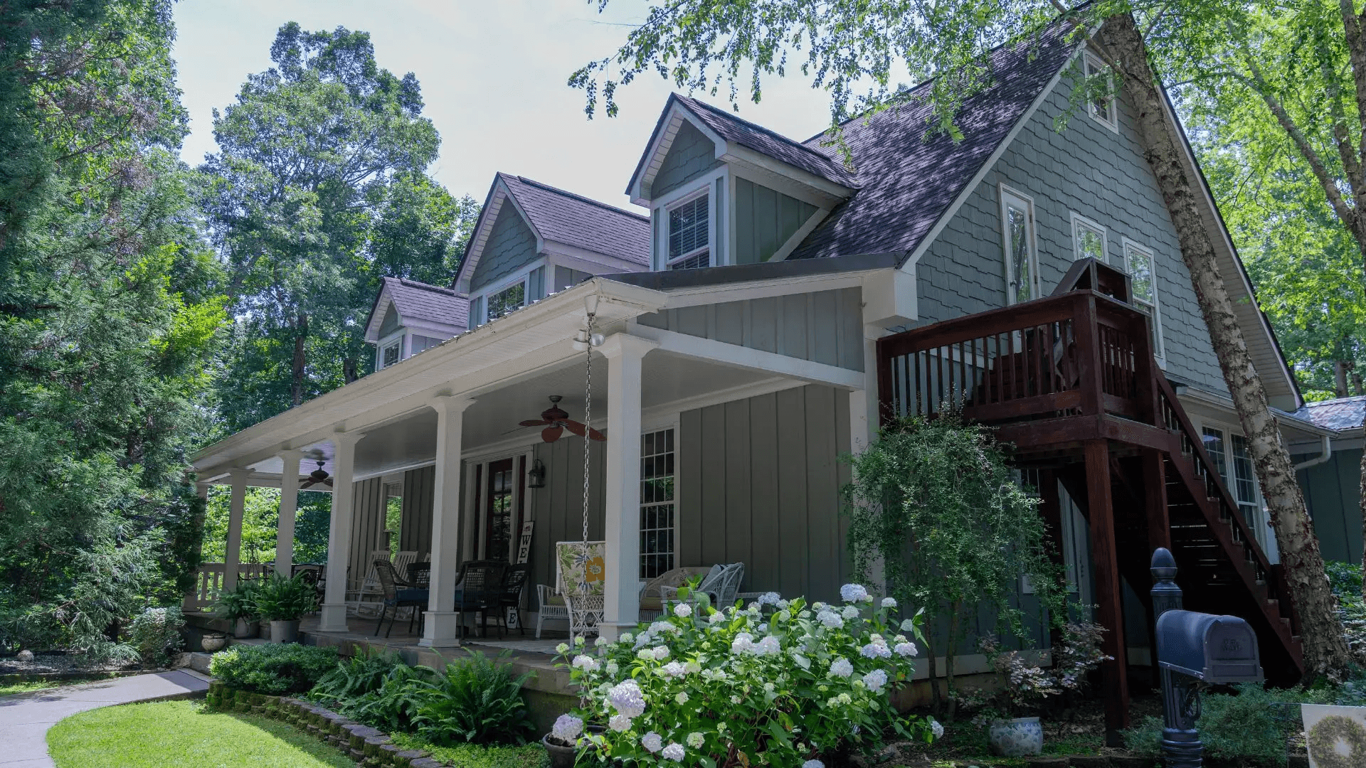Charming house with light green siding, a spacious wraparound porch, white columns, lush greenery, white flowers, and a peaceful, inviting atmosphere.