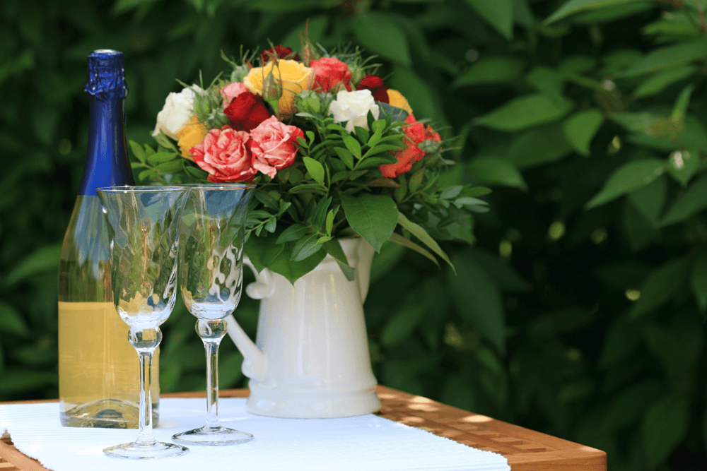 A bottle of sparkling wine with two glasses and a colorful bouquet of flowers on a wooden table.