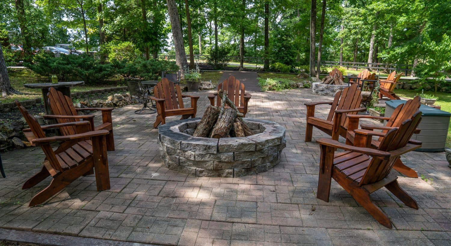 A circular stone fire pit surrounded by wooden Adirondack chairs in a wooded outdoor setting.