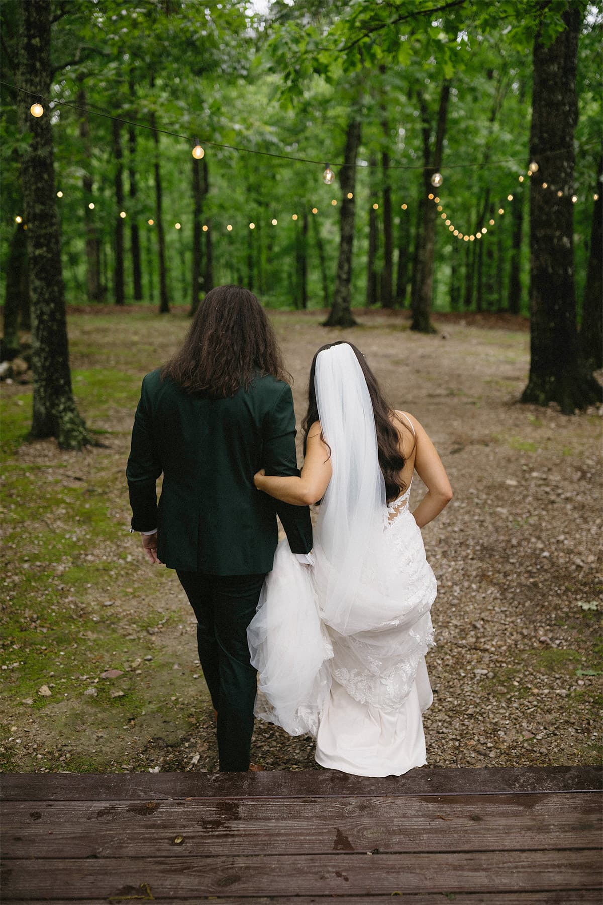 A couple walks hand in hand down a pathway in a lush green forest, adorned with string lights. A couple walks hand in hand down a pathway in a lush green forest, adorned with string lights.