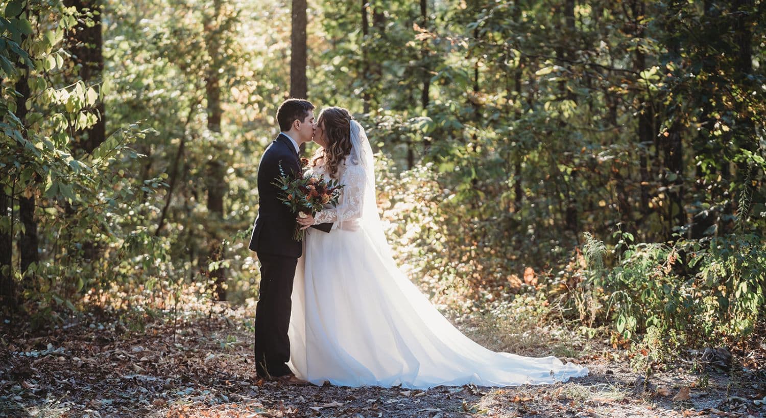 A bride and groom share a kiss in a sunlit forest.