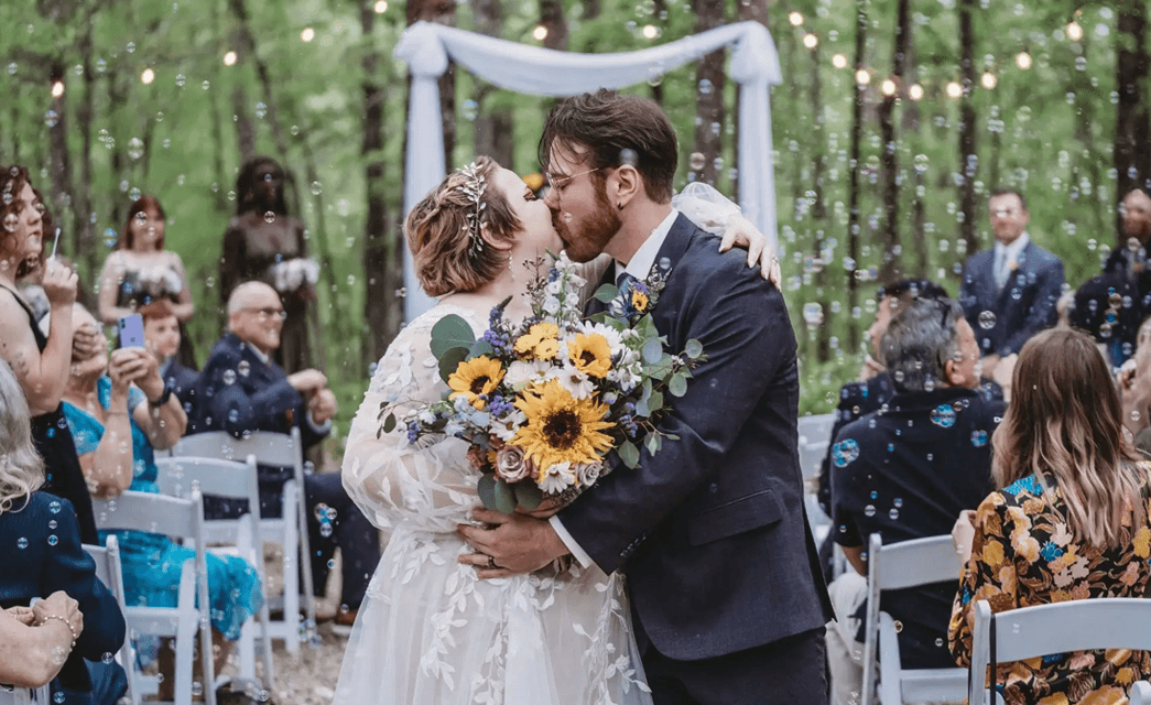 A bride and groom kiss during their outdoor wedding ceremony surrounded by guests and floating bubbles.