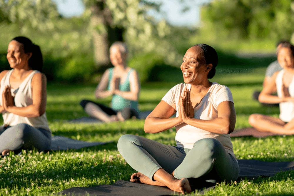 A group of people practicing yoga outdoors, sitting cross-legged with hands in a prayer position and smiling.
