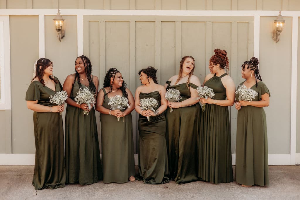 A group of seven bridesmaids in olive green dresses, each holding small bouquets of flowers, stand together smiling in front of a light green wall.