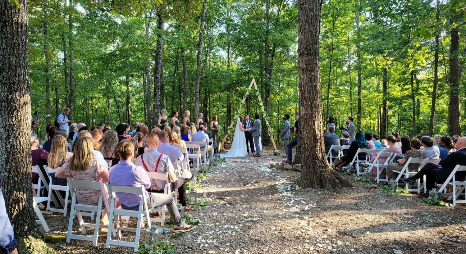 An outdoor wedding ceremony takes place in a forest with guests seated on either side.