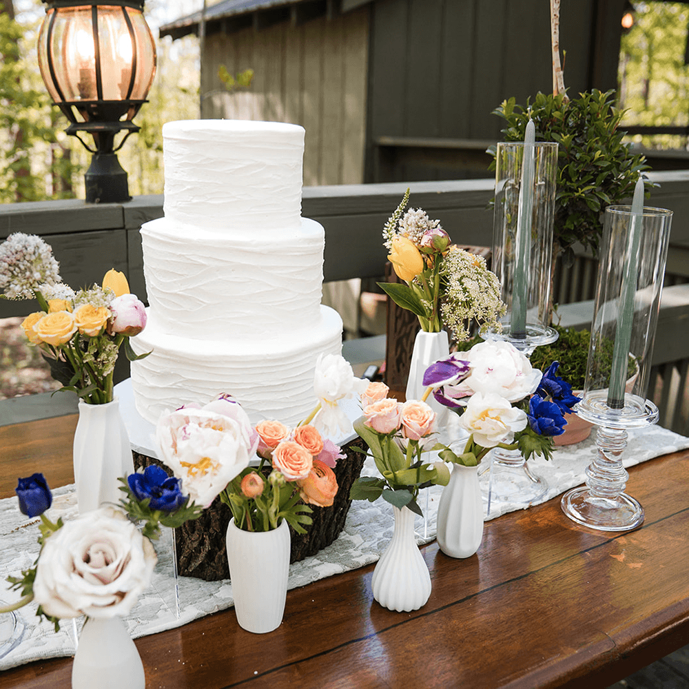 A three-tiered white wedding cake is beautifully adorned with floral arrangements on a wooden table.
