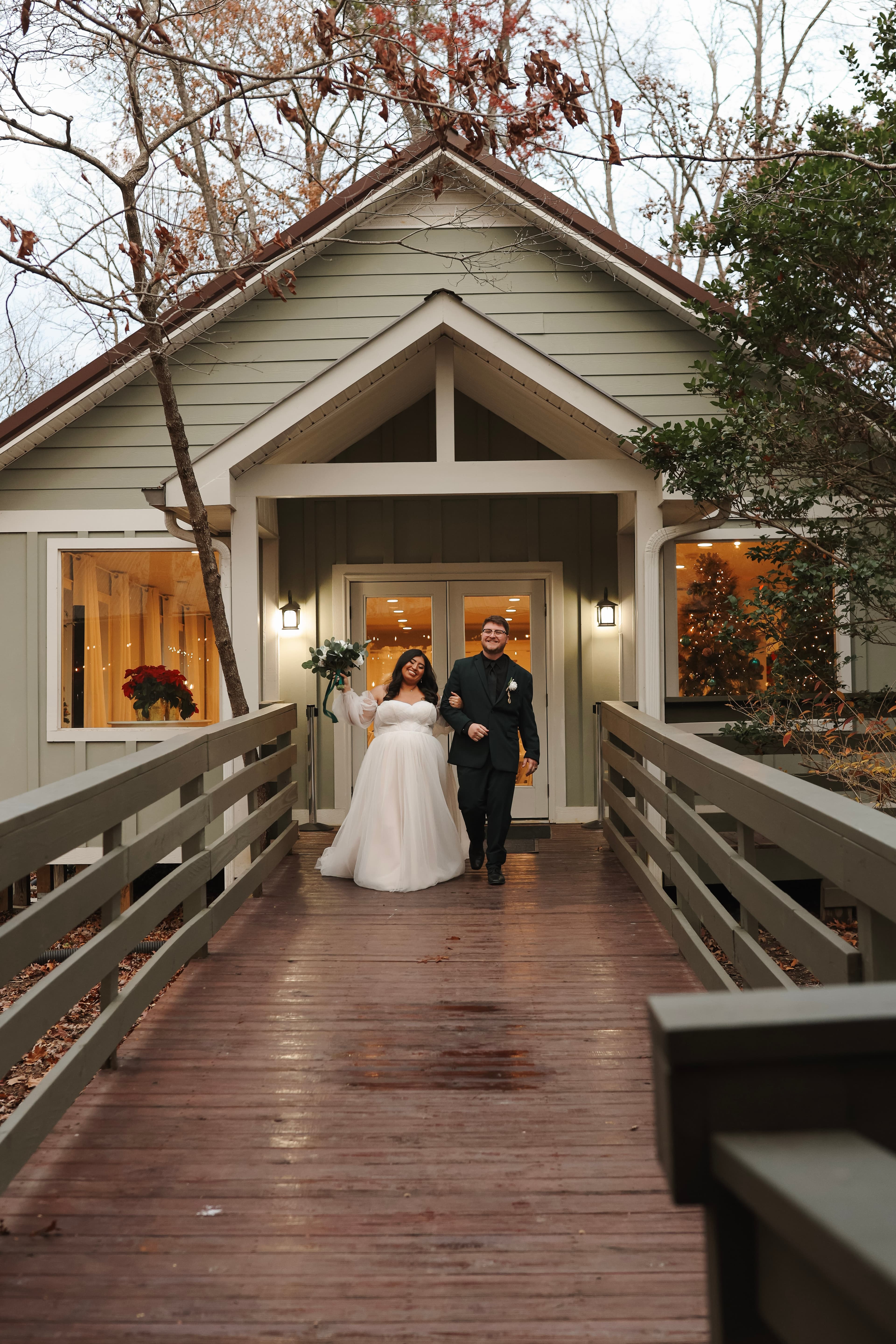 A bride and groom joyfully walk together down a wooden path toward a cozy, decorated house surrounded by trees.