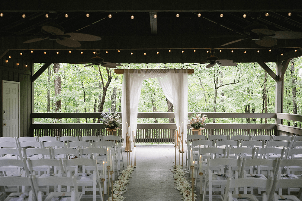 An outdoor wedding venue featuring white chairs arranged in rows facing a wooden altar adorned with sheer drapes. Floral decorations and trees in the background.