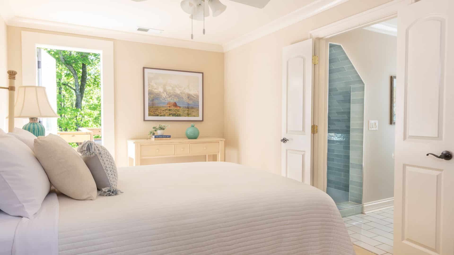A bright and inviting bedroom featuring a large bed, a dresser, and an open door leading to a patio with greenery.
