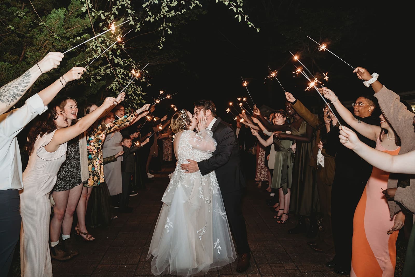 A newlywed couple kisses while guests celebrate with sparklers in a night setting. A newlywed couple kisses while guests celebrate with sparklers in a night setting.