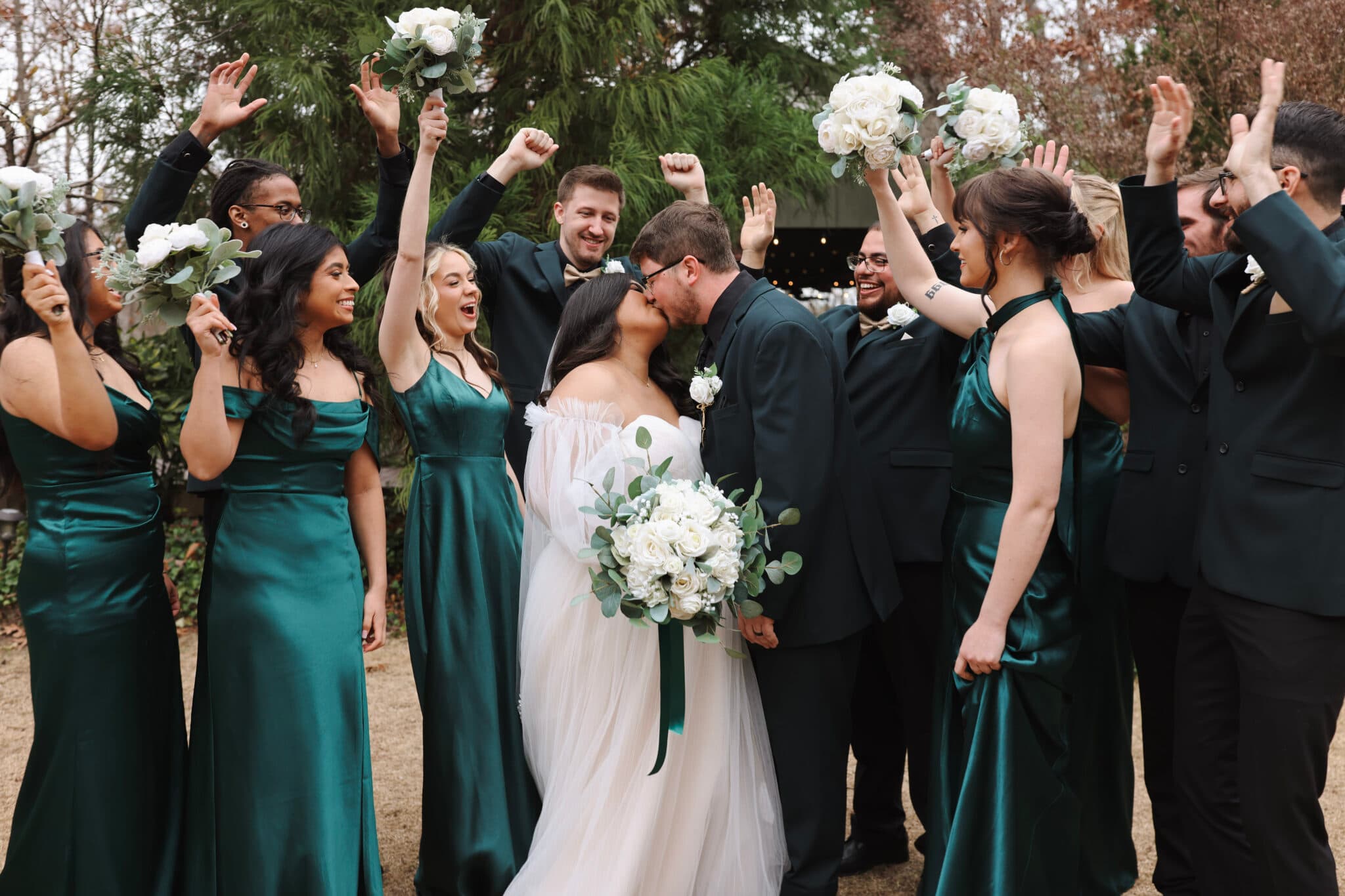 A bride and groom kiss surrounded by their joyful bridal party, all celebrating outdoors. A bride and groom kiss surrounded by their joyful bridal party, all celebrating outdoors.
