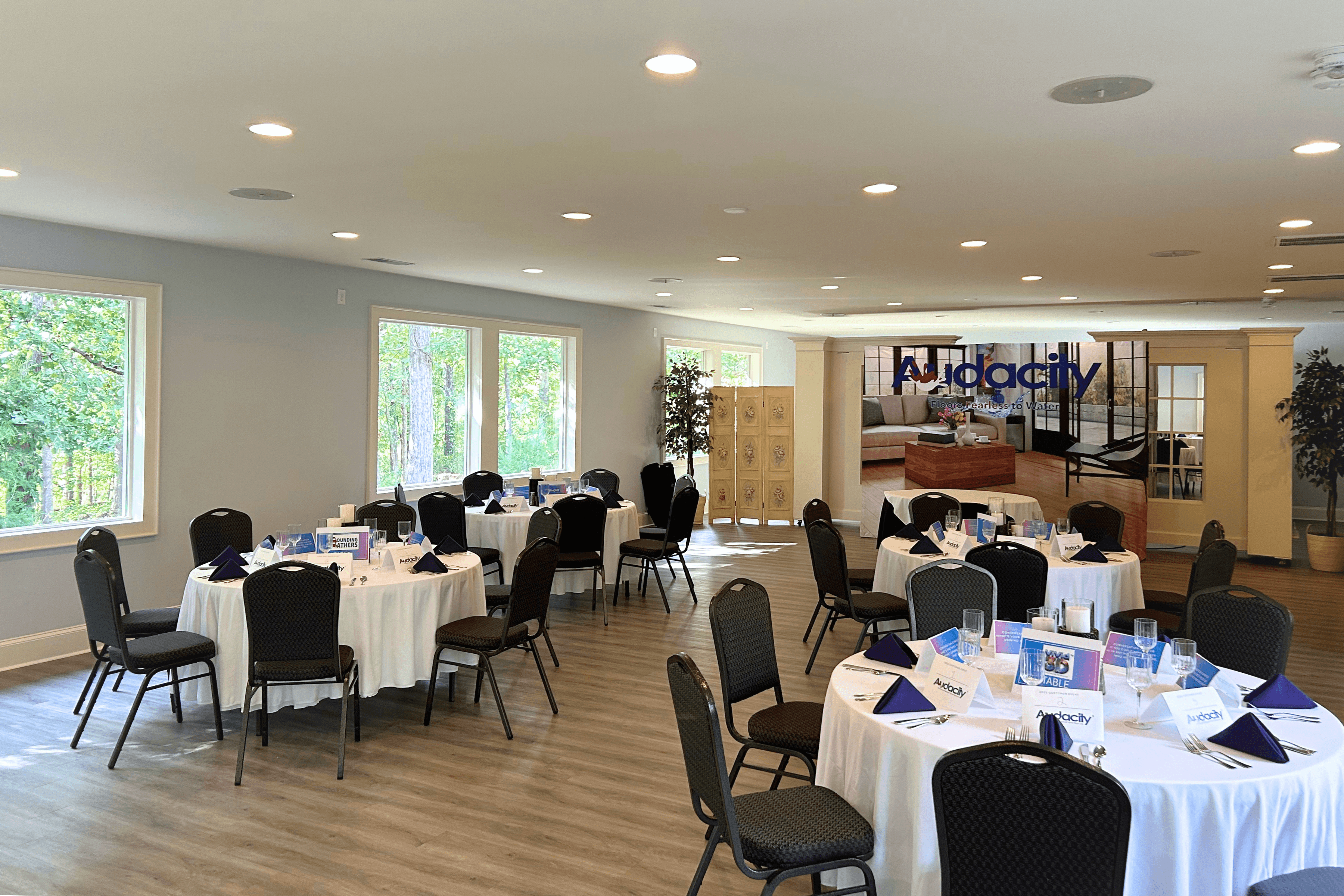 Bright event room with round tables set for a meeting, featuring white tablecloths, black chairs, folded navy napkins, and Audacity-branded materials, with large windows overlooking trees