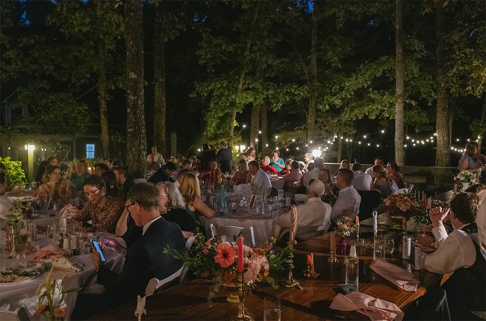 A nighttime outdoor gathering with people seated at tables, illuminated by string lights. The atmosphere is warm and festive, surrounded by trees.
