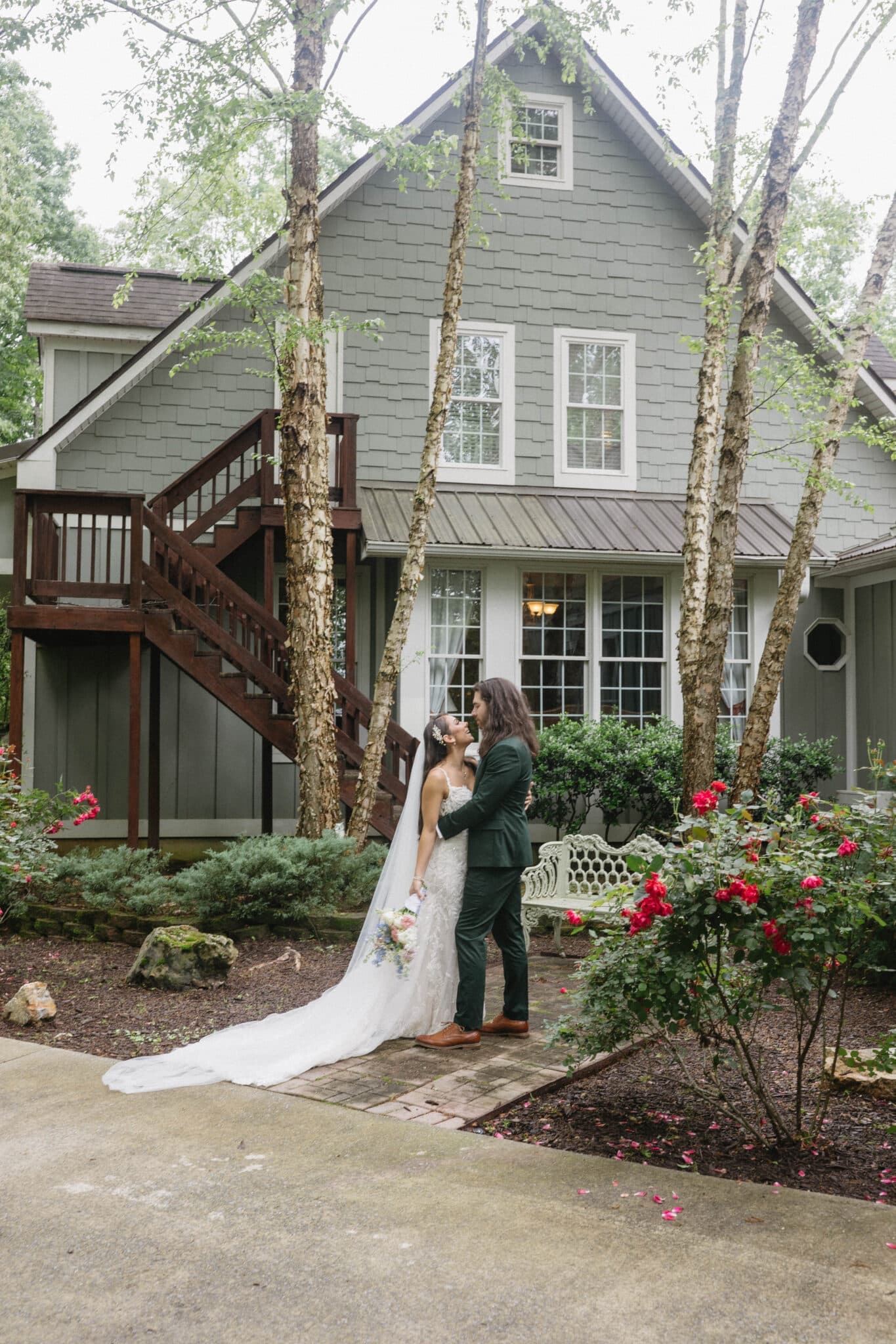 A bride and groom share a kiss in front of a gray house surrounded by trees and blooming flowers. A bride and groom share a kiss in front of a gray house surrounded by trees and blooming flowers.