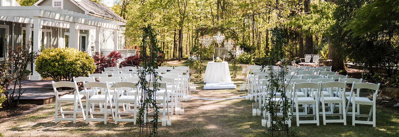 Outdoor wedding ceremony setup with rows of white chairs facing an altar under a lush canopy of trees. A charming white house is visible on the left.