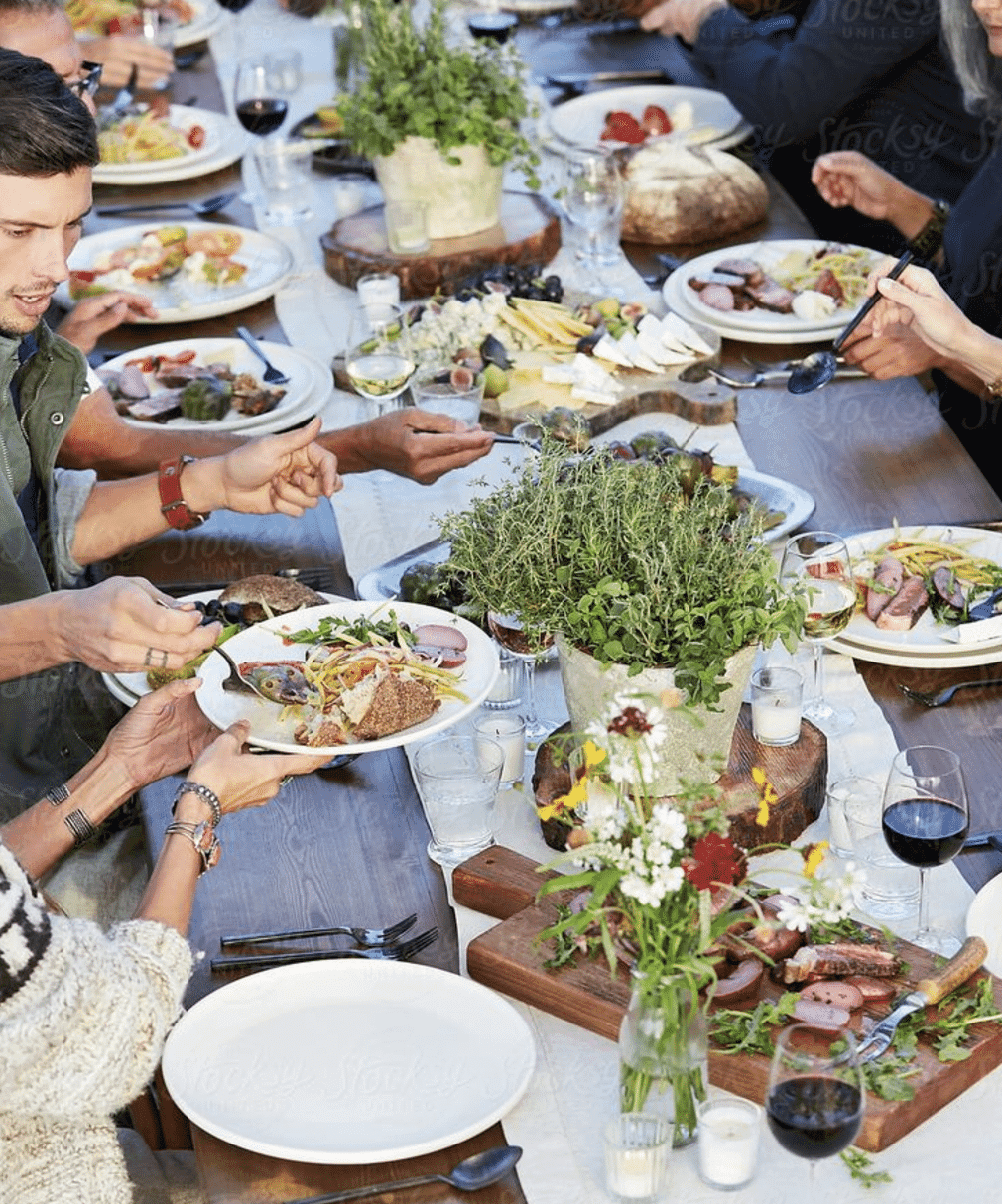 A vibrant outdoor gathering featuring people enjoying a variety of dishes and drinks at a long table adorned with fresh herbs and flowers. A vibrant outdoor gathering featuring people enjoying a variety of dishes and drinks at a long table adorned with fresh herbs and flowers.
