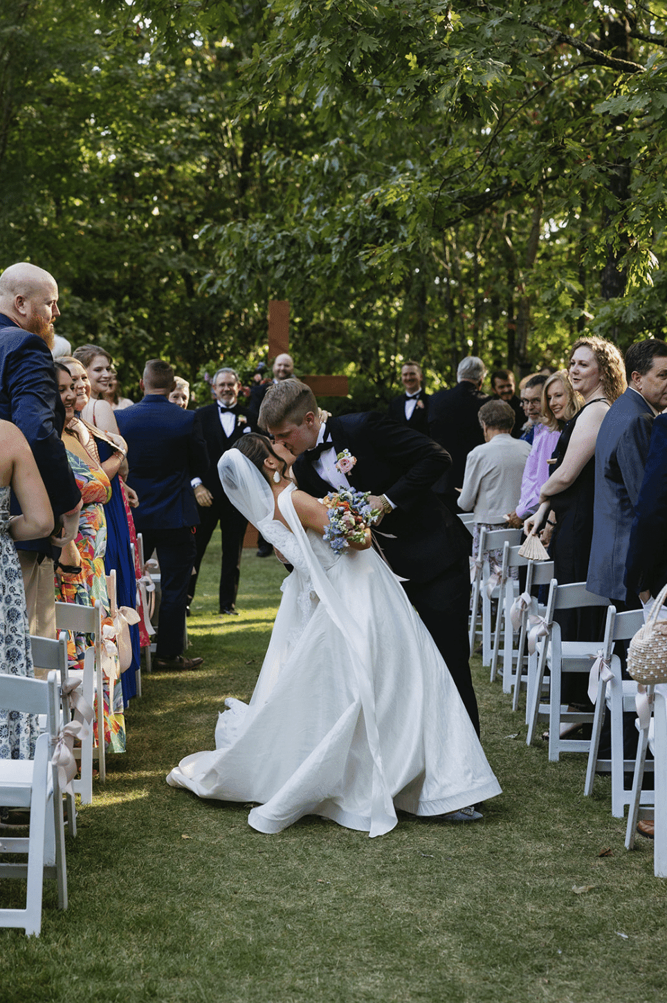 A bride and groom share a kiss during their outdoor wedding ceremony surrounded by guests. A bride and groom share a kiss during their outdoor wedding ceremony surrounded by guests.