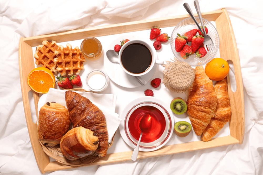 A wooden tray filled with croissants, waffles, fresh fruit, honey, coffee, and tea.