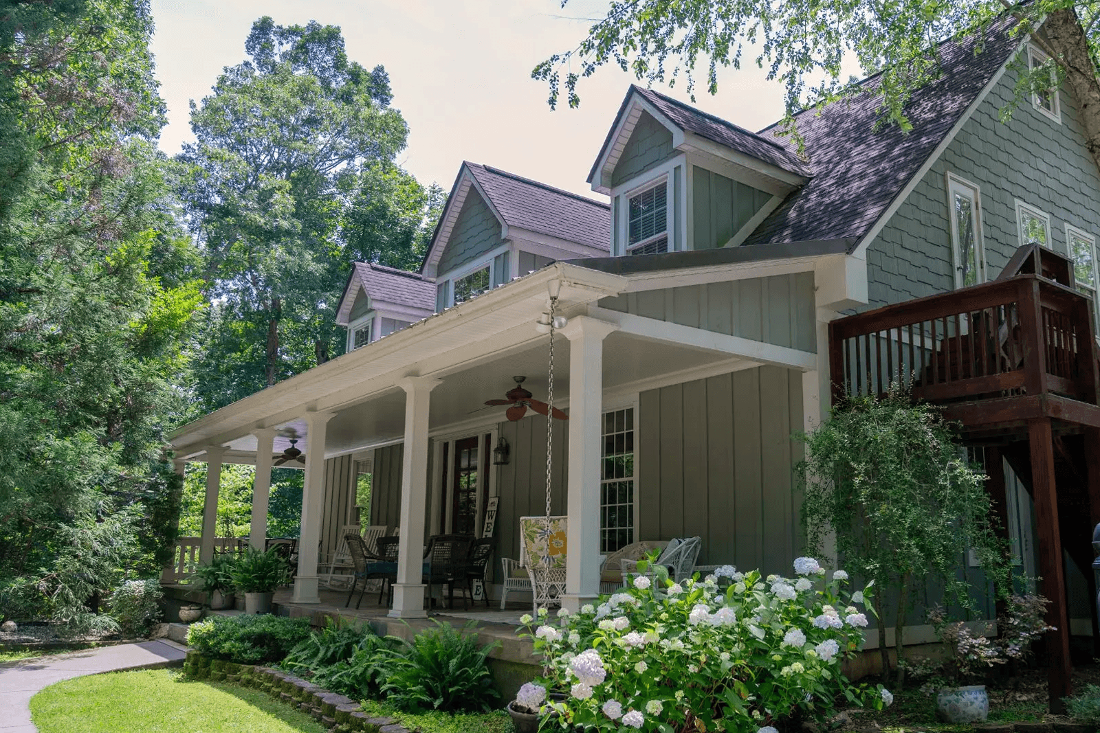 A cozy green house with a porch, surrounded by trees and flowering plants.