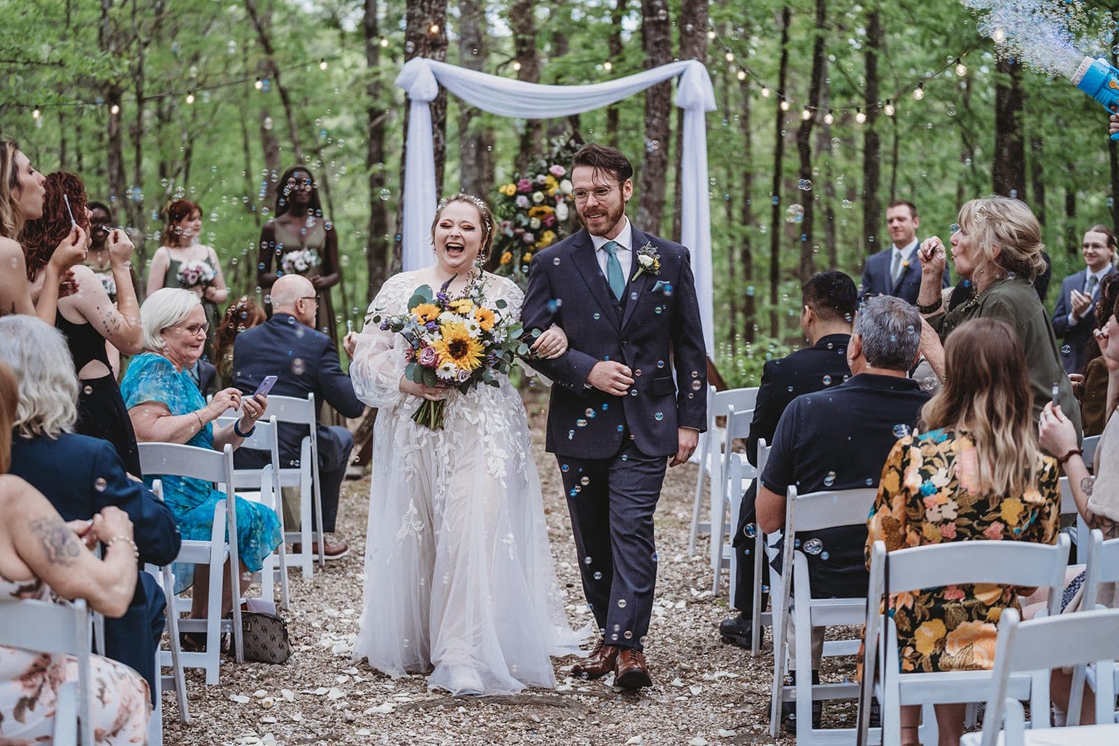 A smiling bride and groom walk down the aisle surrounded by guests and bubbles in a forest setting. A smiling bride and groom walk down the aisle surrounded by guests and bubbles in a forest setting.