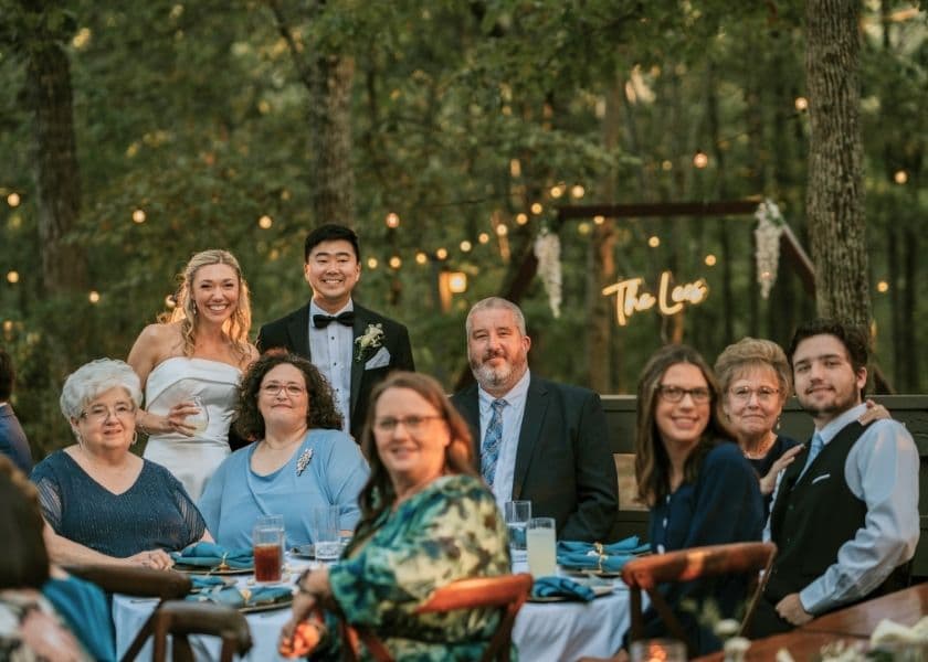 A joyful group of guests, including a bride and groom, gather around a table decorated with blue accents in a forest setting.