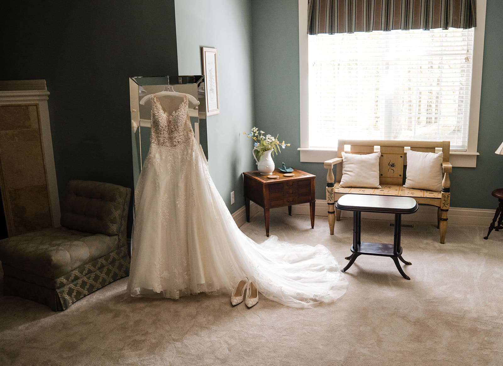 A wedding dress hangs on a mirror in a softly lit room with a vintage chair, a wooden table, and floral decor.