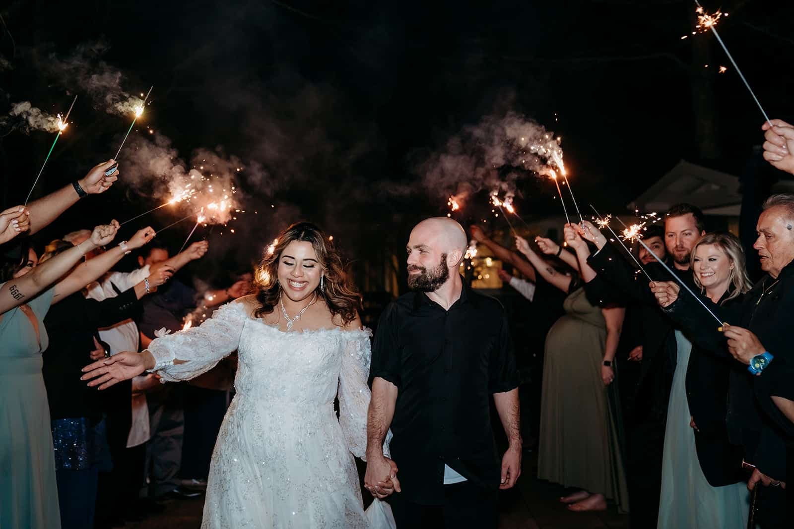 A bride and groom joyfully walk hand-in-hand through a crowd holding sparklers on a festive night.