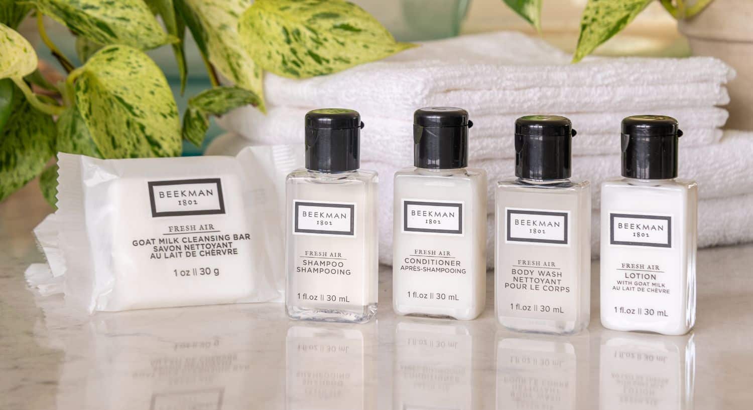 Four Beekman 1802 skincare products, including a cleansing bar, shampoo, conditioner, body wash, and lotion, on a countertop with greenery and towels.