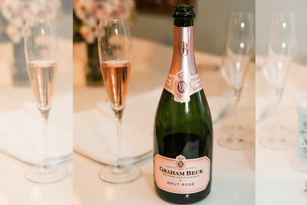 A bottle of Graham Beck Brut Rosé sits on a table next to two fluted glasses filled with sparkling rosé.
