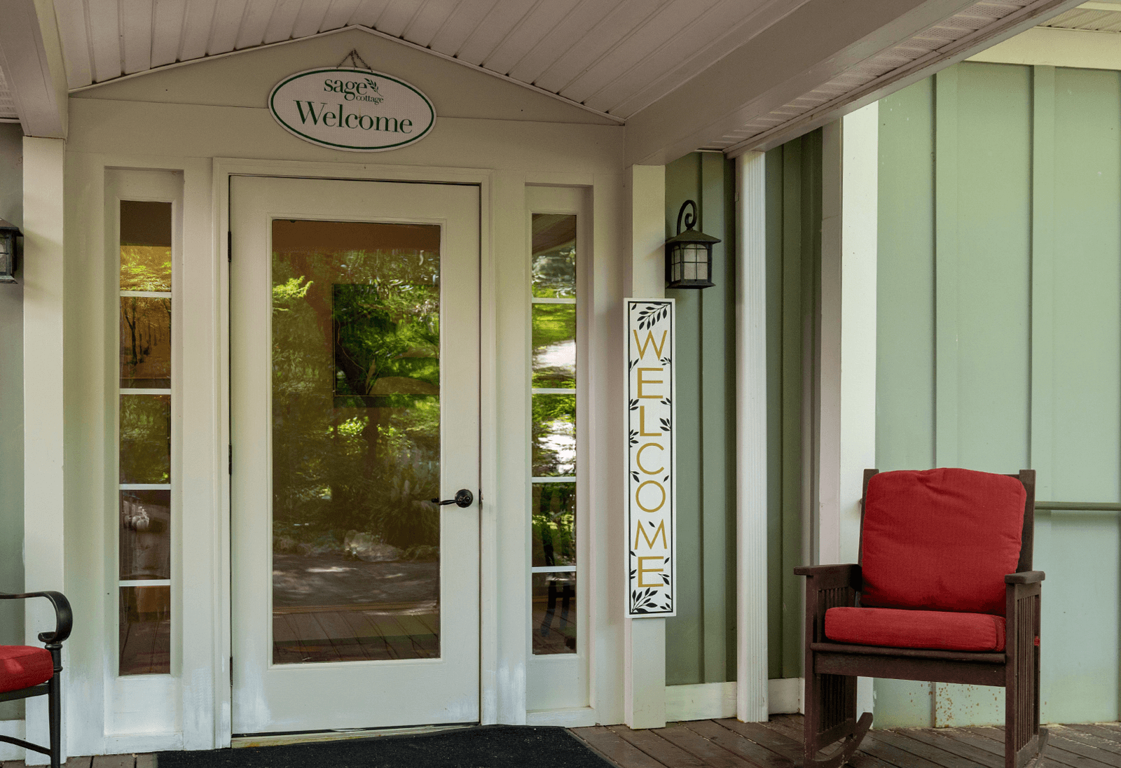 Cream-colored double doors with glass panels are flanked by welcome signs. A red cushioned chair sits on the wooden porch, creating a cozy, inviting entrance.