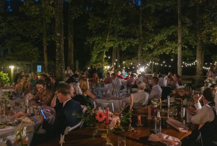 A lively outdoor gathering at night, with tables filled with guests, string lights, and floral decorations. A lively outdoor gathering at night, with tables filled with guests, string lights, and floral decorations.